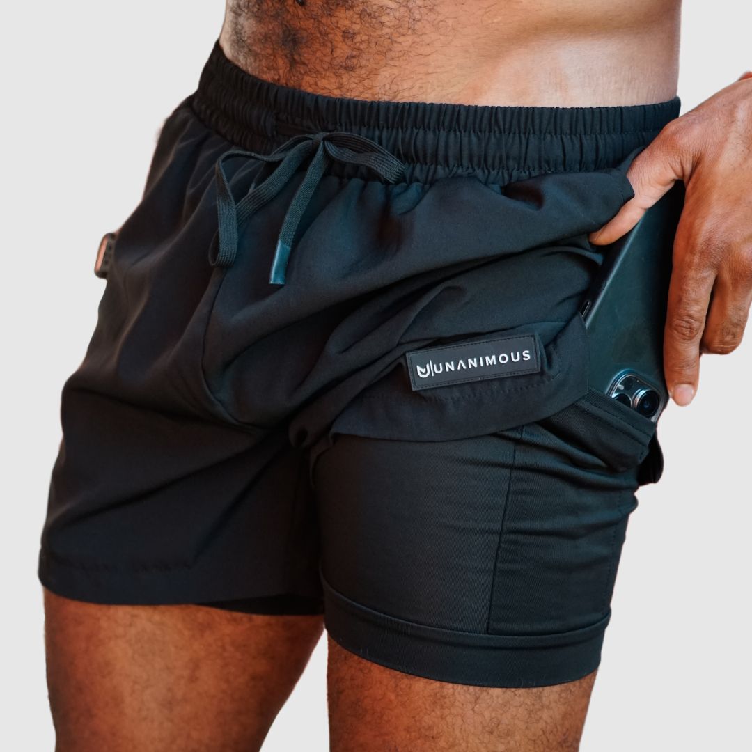 Black 2-in-1 men's training shorts, model showing phone in inner compression pocket.