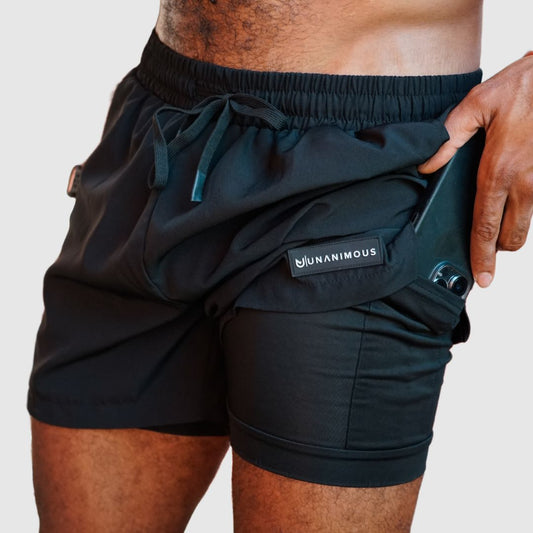 Black 2-in-1 men's training shorts, model showing phone in inner compression pocket.