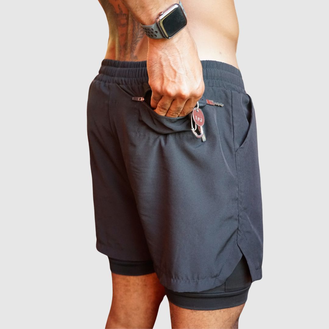 Black 2-in-1 men's athletic shorts, model reaching into side storage pocket.